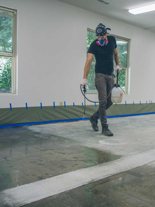 Person applying liquid to a floor using a spray gun in a room with windows and blue tape on the floor.