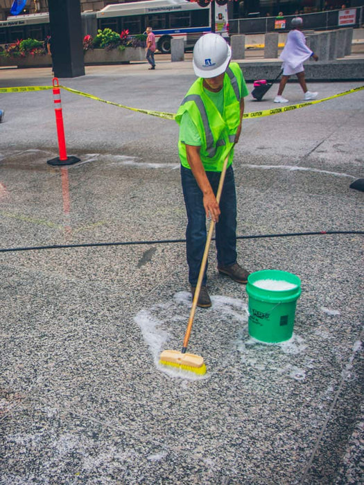 Person cleaning a street with a broom and bucket, with a bus and people in the background.