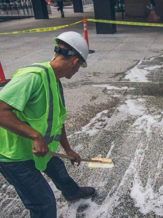 Person in high-visibility vest and hard hat using a tool on a road surface.