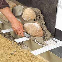 A person's hand placing a stone into a cavity on a wall, with mortar and tools visible on the ground.