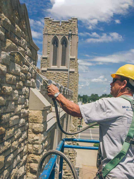 Person using a pressure washer on a stone building with a church tower in the background